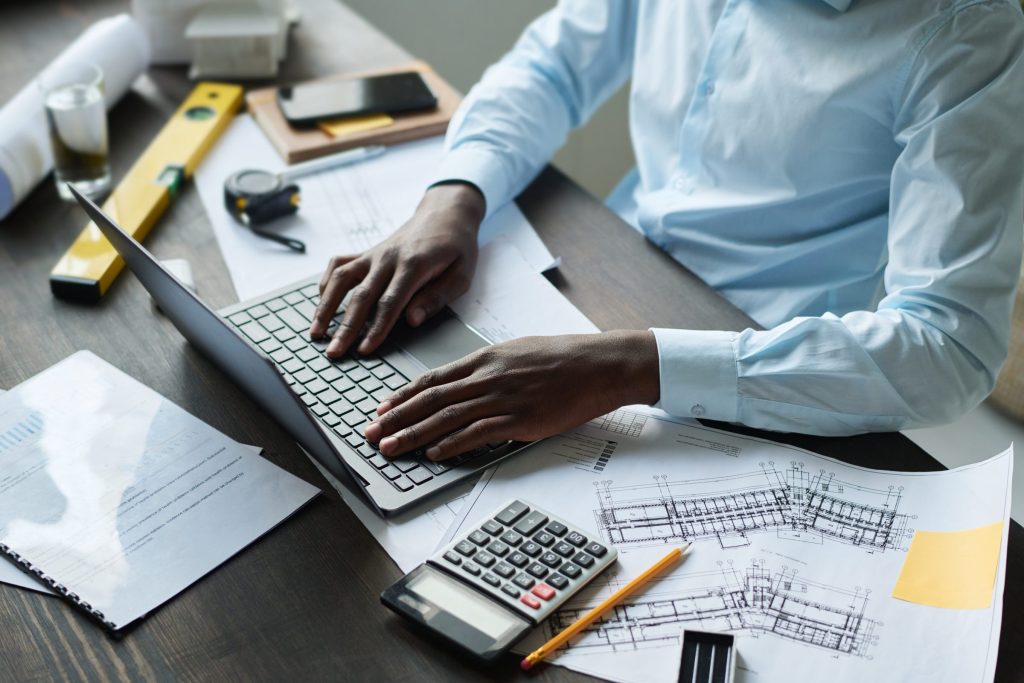 Hands at a laptop on a desk with a calculator, a tape measure and drawings, working out a figure