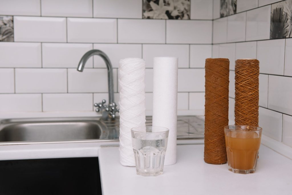 New and spent water filter cartridges beside a glass of clear water and a glass of discolored water