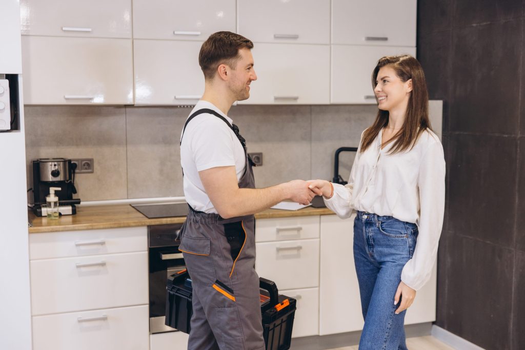 A tradesman in overalls shaking hands with a homeowner in a kitchen, tool case at his feet