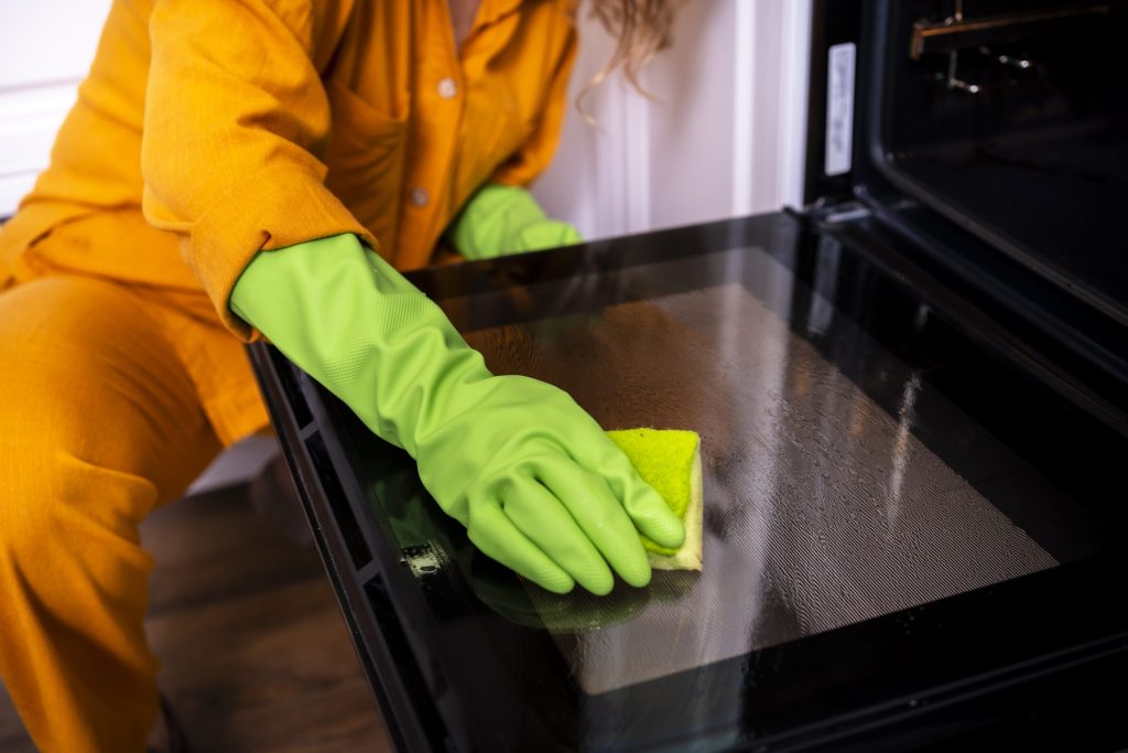 A gloved hand wiping the inside of a black oven door with a sponge