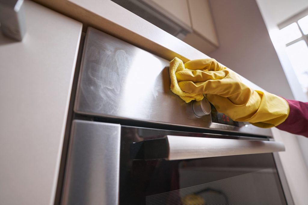 A gloved hand wiping the stainless handle and glass of a wall oven