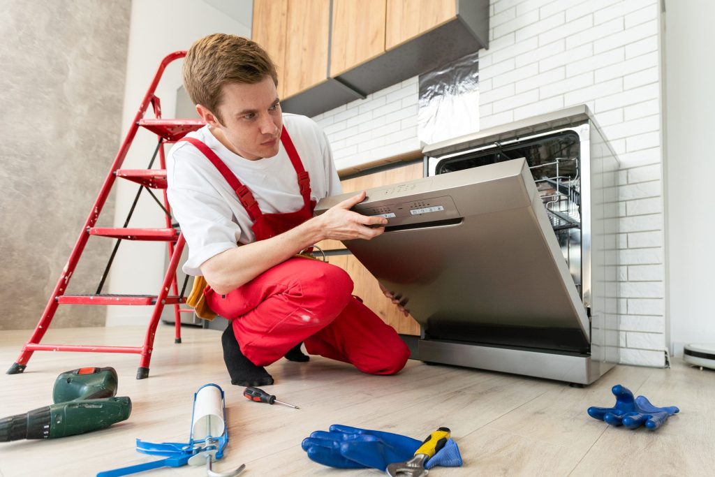 A technician kneeling at an open dishwasher door with tools around him