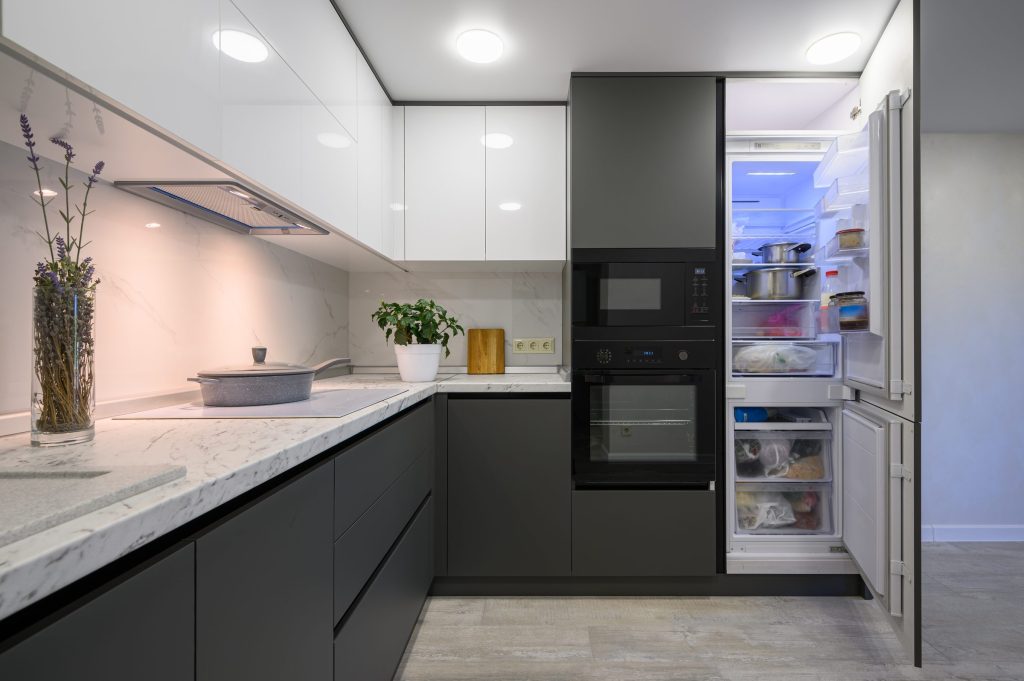 A grey kitchen with a built-in oven and microwave column beside an open refrigerator