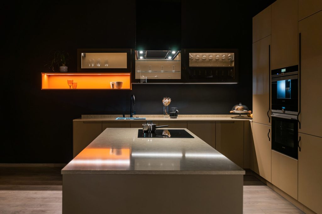 A dark contemporary kitchen lit by warm strip lighting, with a steel island and a tall oven column