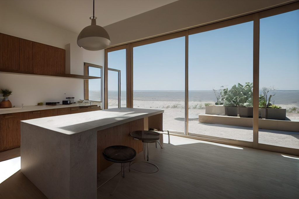 A concrete island with stools in a walnut kitchen opening onto a dune terrace