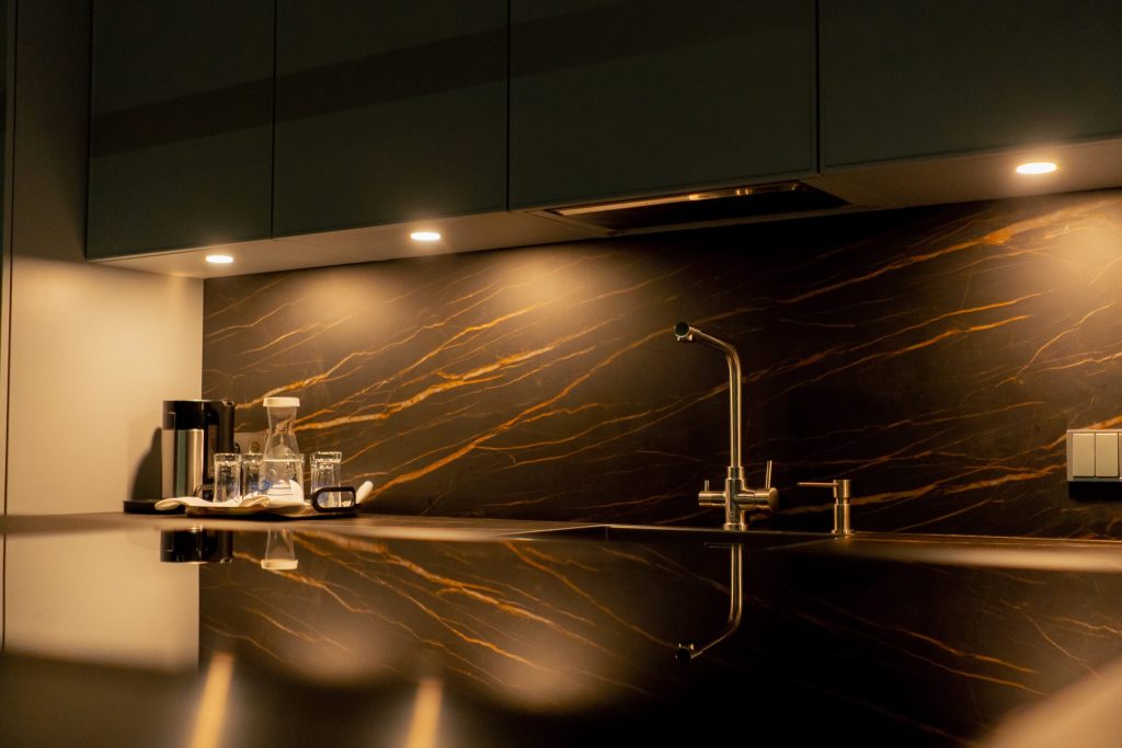 A dark kitchen counter against a gold-veined stone splashback, with a brass tap and glassware