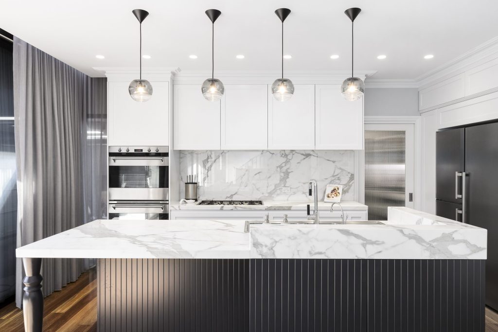 A white kitchen with a marble island and splashback, built-in ovens and a graphite refrigerator