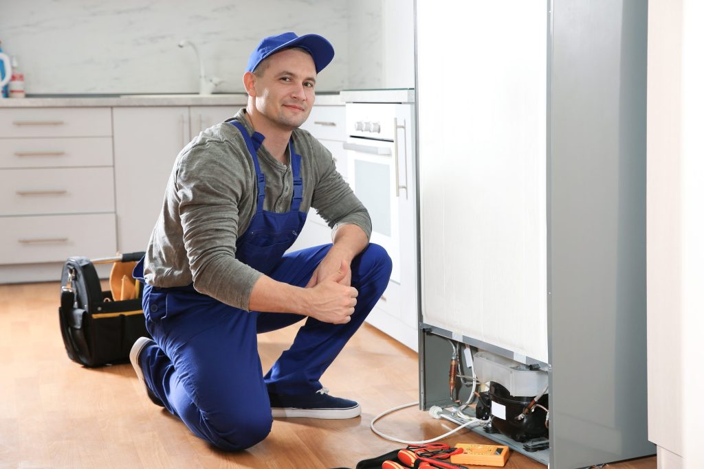 A technician kneeling beside a refrigerator with the kickplate off and his tools on the floor