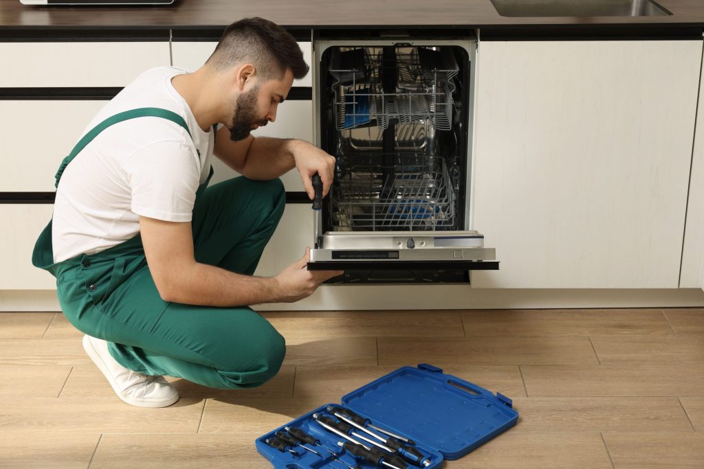 A technician kneeling at an open built-in dishwasher, working on the door with a screwdriver, tool case on the floor