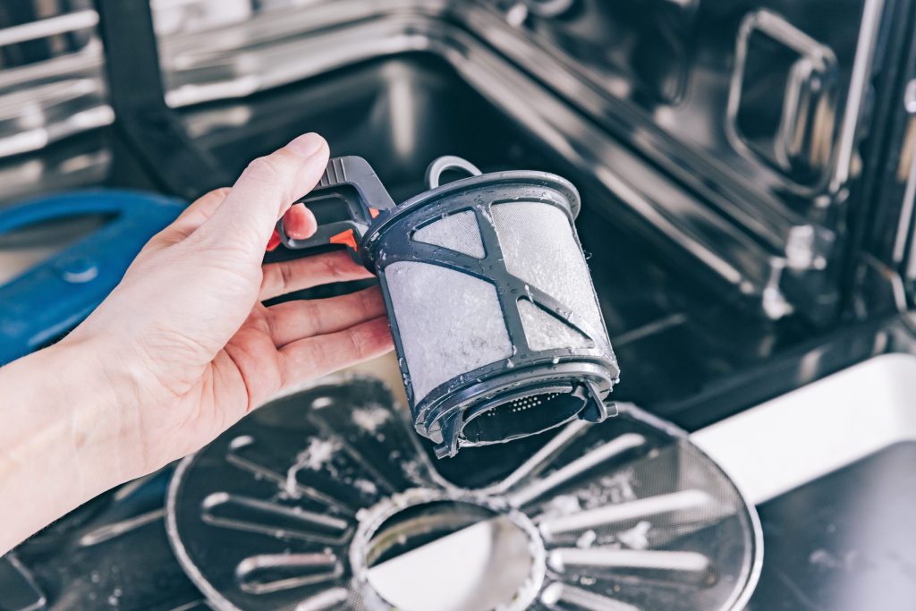 A hand lifting the cylindrical filter out of the sump of an open dishwasher, limescale on the mesh