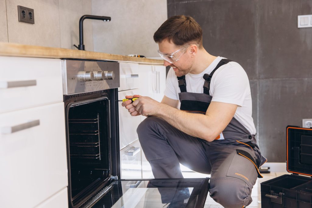 A fitter in safety glasses kneeling at a built-in wall oven, driving a screw into the cabinet frame