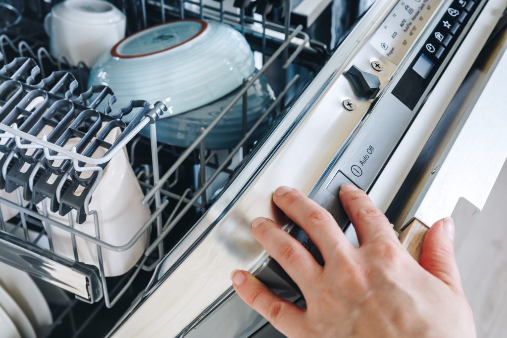 A hand pressing the start control on the top edge of an open dishwasher door
