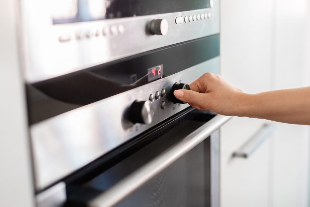 A hand turning the temperature dial on a stainless wall oven with a red display