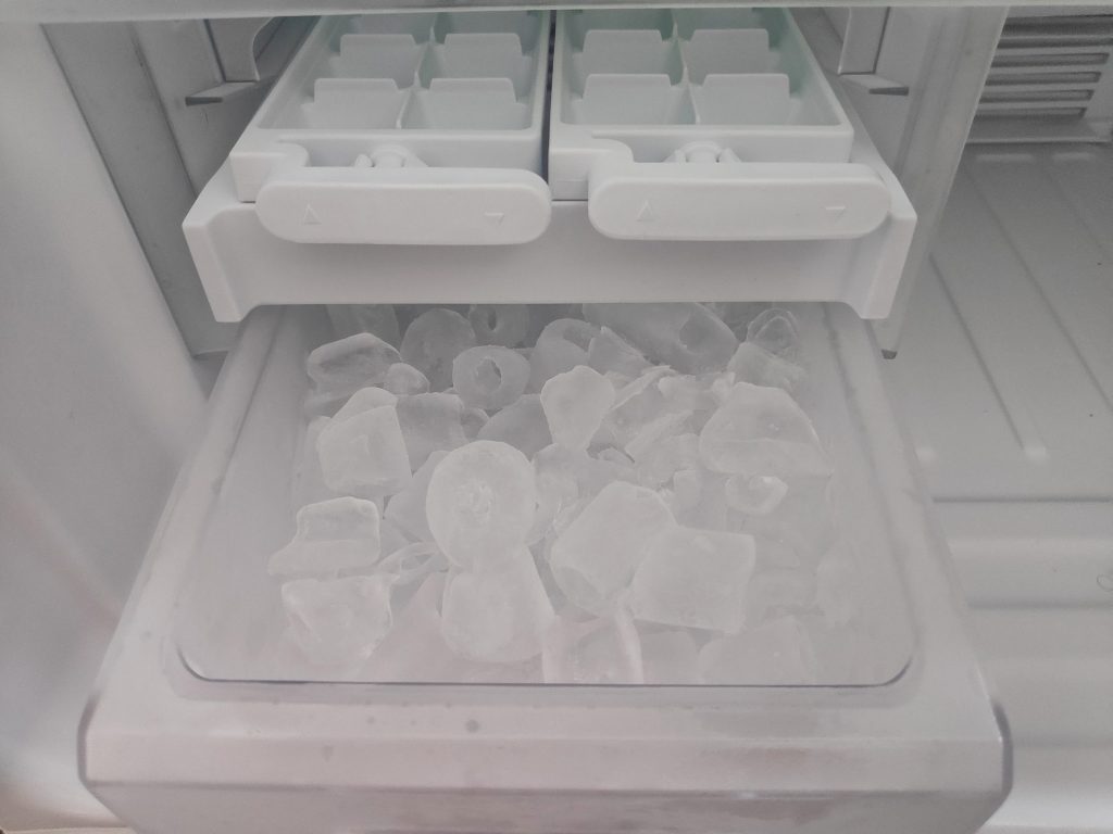 Ice cubes filling the bin below two ice trays inside a freezer